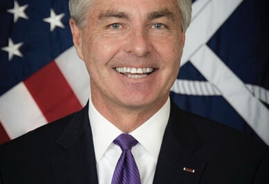 A man with gray hair in a dark suit, white shirt, and purple tie smiles in front of an American flag and another flag with a white emblem.
