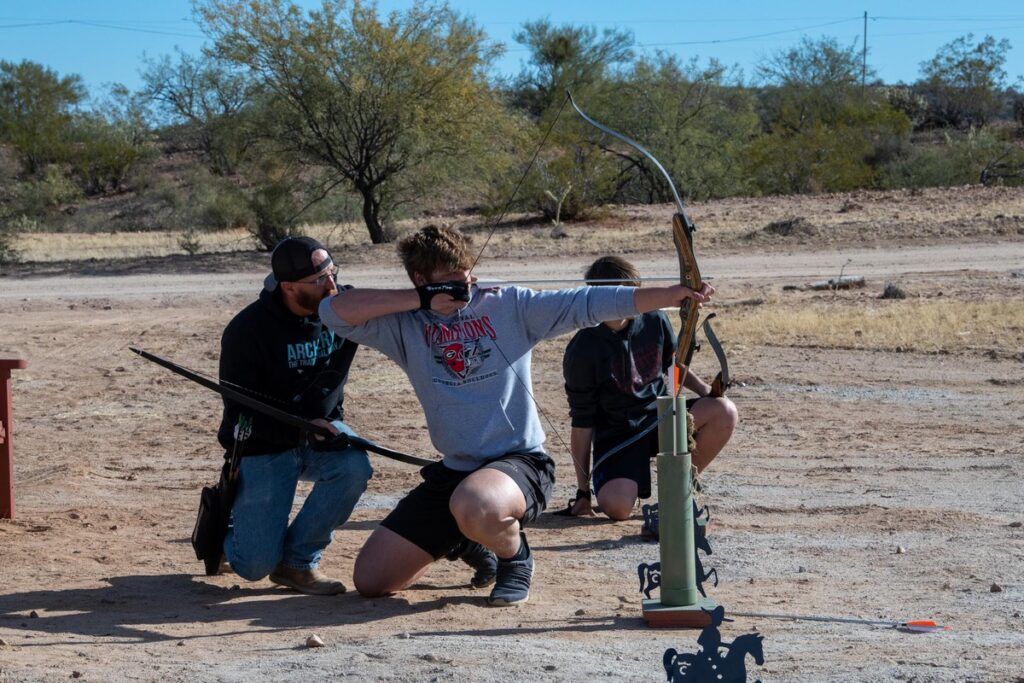 Three people are outdoors on a sandy range practicing archery. One person kneels, aiming a bow, while another kneels beside them and a third person stands, appearing to instruct or observe. Desert vegetation is in the background.