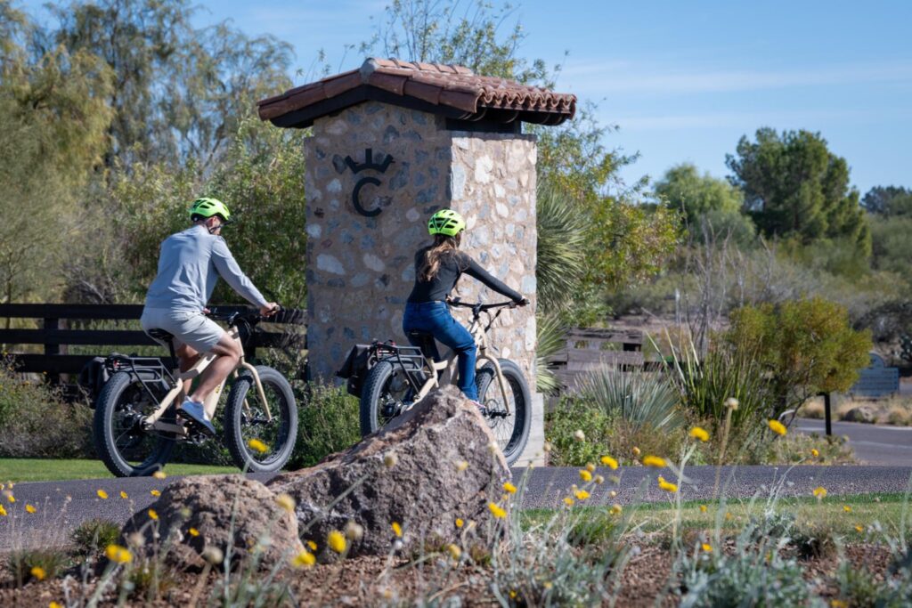 Two people wearing helmets ride electric bikes past a stone entrance sign with a "C" and a crown symbol, surrounded by desert plants, rocks, and a paved road under a clear blue sky.