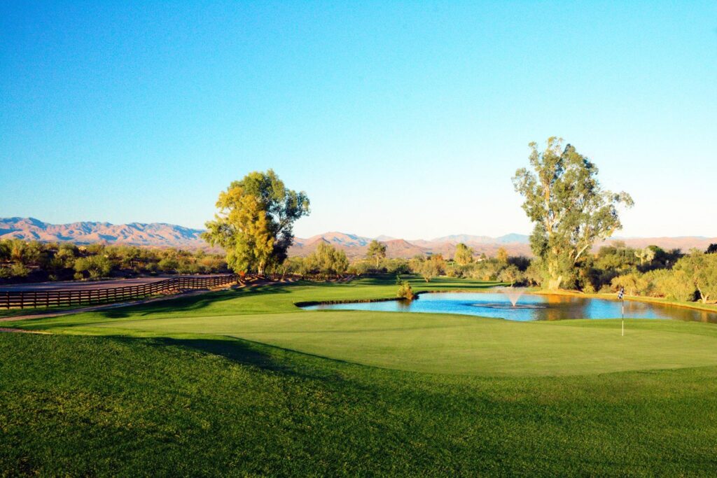 A bright, sunny day on a golf course with lush green grass, a small pond, tall trees, wooden fencing, and distant mountains under a clear blue sky.
