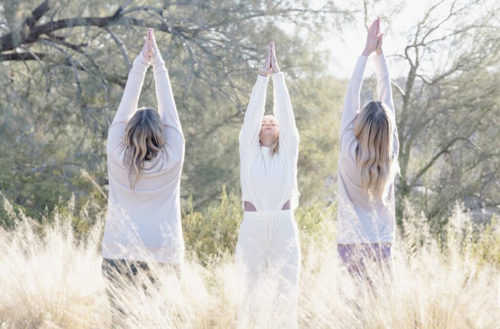 Three women stand outdoors in tall grass, facing away with arms raised and palms together in a yoga pose. The scene is bright and peaceful, with sunlight filtering through trees in the background.