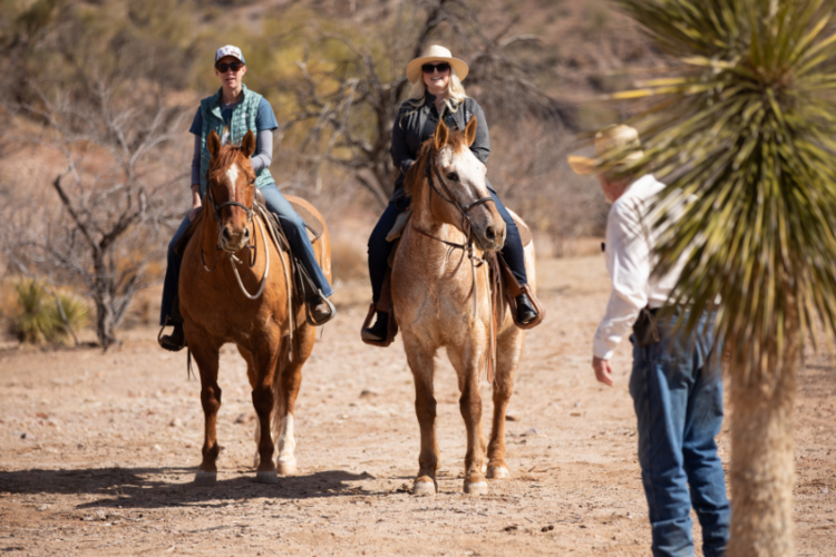 Two people on horseback, both wearing hats and sunglasses, ride along a dry, desert trail with sparse vegetation. A third person, also wearing a hat, stands in the foreground facing them.