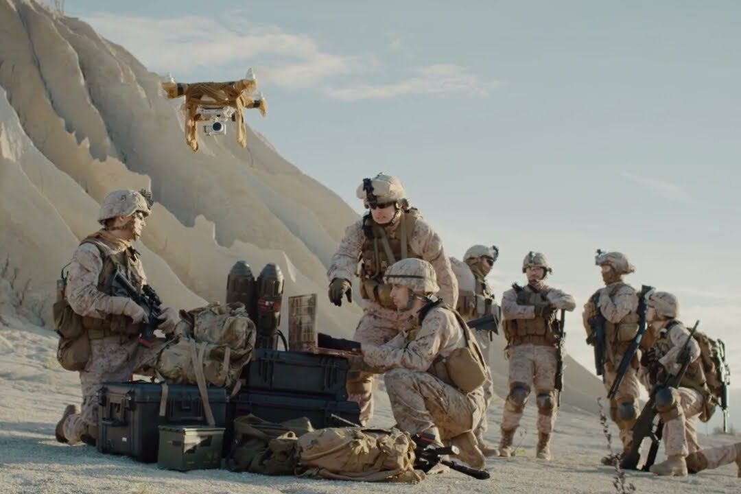 A group of soldiers in desert camouflage gear operate equipment and launch a drone in a rocky, arid landscape, while others stand in the background observing.