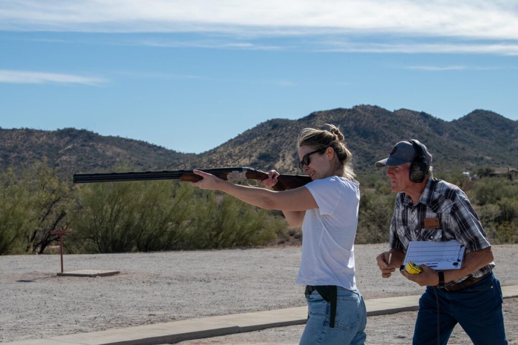 A woman aiming a shotgun at an outdoor shooting range, assisted by an older man wearing ear protection and holding a clipboard, with desert hills and clear skies in the background.