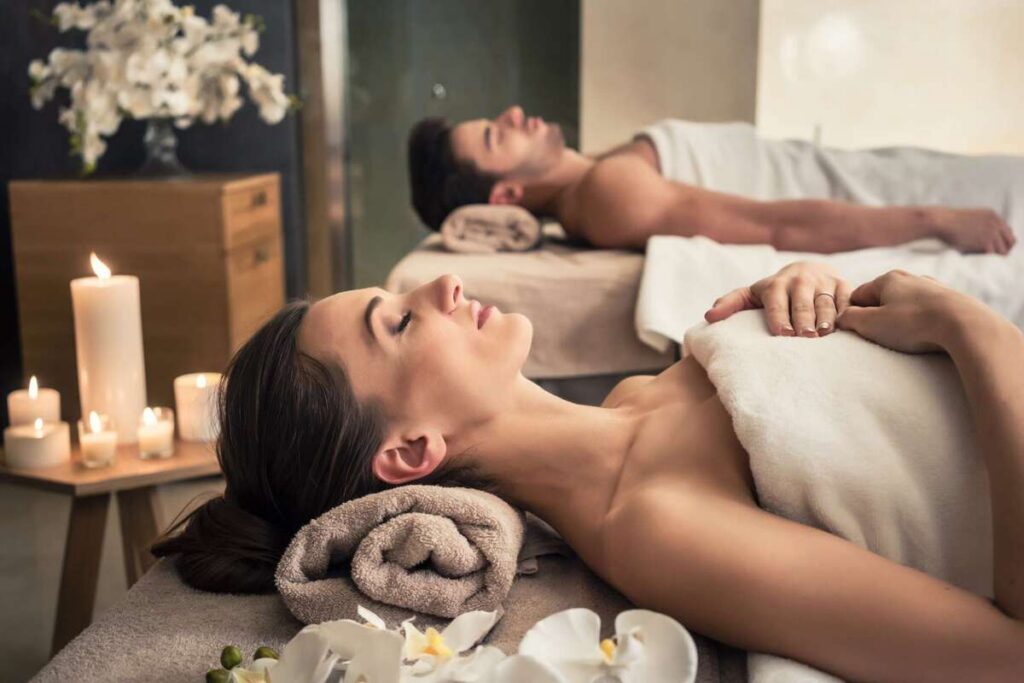 A woman and a man relax on separate massage tables with towels and blankets in a serene spa setting, surrounded by lit candles, flowers, and soft lighting.