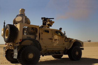 A tan military armored vehicle equipped with a mounted gun drives across a flat, sandy desert landscape under a clear blue sky. A projectile is visible in mid-air near the front of the vehicle.