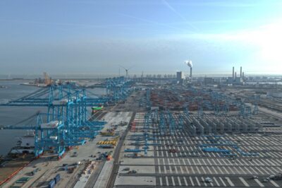 Aerial view of a large industrial port with blue cranes, shipping containers, and warehouses along the waterfront. Factories and wind turbines are visible in the background under a clear sky.