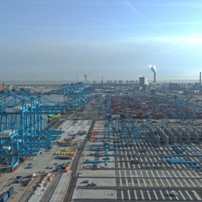 Aerial view of a large industrial port with blue cranes, shipping containers, and warehouses along the waterfront. Factories and wind turbines are visible in the background under a clear sky.