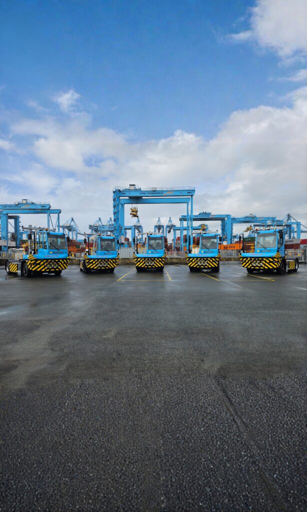 Five blue cargo trucks lined up side by side on a wet asphalt surface at a shipping port, with cranes, shipping containers, and a partly cloudy sky in the background.