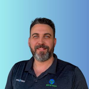 A man with short dark hair and a beard, wearing a black collared shirt with “BRITE FORCE” and “BRITE TECH” logos, smiling in front of a blue gradient background.