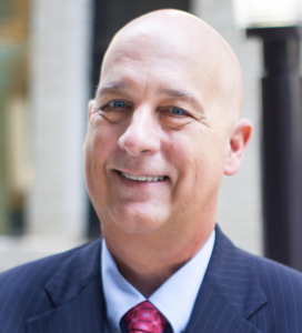 A smiling middle-aged man in a dark pinstripe suit, white shirt, and red tie stands outdoors in front of a blurred urban background. He is bald and has blue eyes.