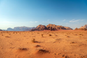 A vast desert landscape with reddish-orange sand, sparse vegetation, and rocky mountains under a clear blue sky. The scene appears arid and sunlit, evoking a sense of solitude and openness.
