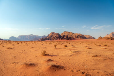 A vast desert landscape with reddish-orange sand, sparse vegetation, and rocky mountains under a clear blue sky. The scene appears arid and sunlit, evoking a sense of solitude and openness.
