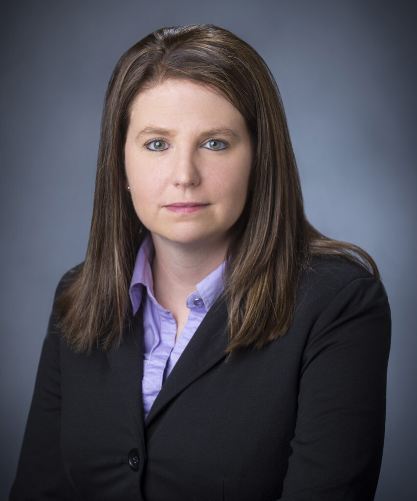 A woman with straight brown hair wearing a black blazer and light purple collared shirt poses for a professional headshot against a plain gray background.