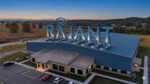 A large industrial building with "RAJANT" in illuminated letters on the roof, surrounded by parking spaces and set in a scenic rural area with trees and hills at sunset.