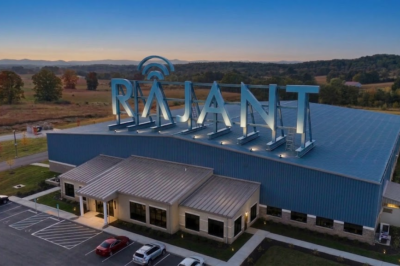 A large industrial building with "RAJANT" in illuminated letters on the roof, surrounded by parking spaces and set in a scenic rural area with trees and hills at sunset.