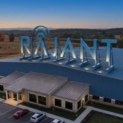 A large industrial building with "RAJANT" in illuminated letters on the roof, surrounded by parking spaces and set in a scenic rural area with trees and hills at sunset.